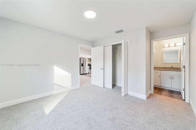 unfurnished bedroom featuring a closet, light colored carpet, stainless steel fridge with ice dispenser, and ensuite bath