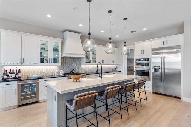 kitchen featuring stainless steel appliances, decorative backsplash, a kitchen island with sink, beverage cooler, and custom range hood