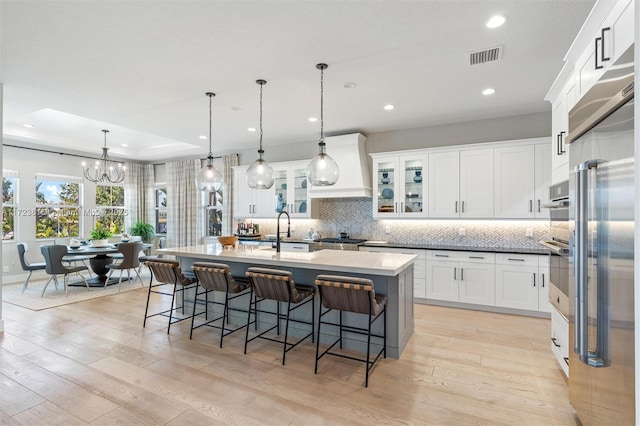 kitchen featuring built in refrigerator, backsplash, a kitchen island with sink, hanging light fixtures, and white cabinets