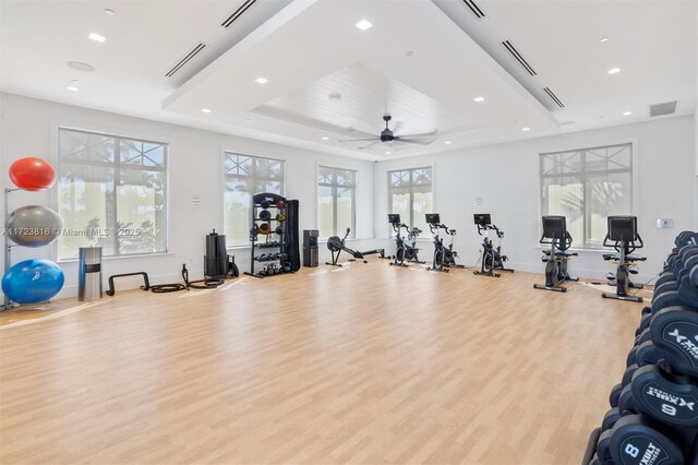 gym with light wood-type flooring, ceiling fan, and a tray ceiling