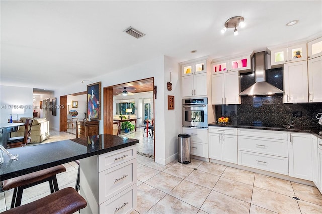 kitchen with backsplash, stainless steel oven, ceiling fan, wall chimney range hood, and white cabinetry