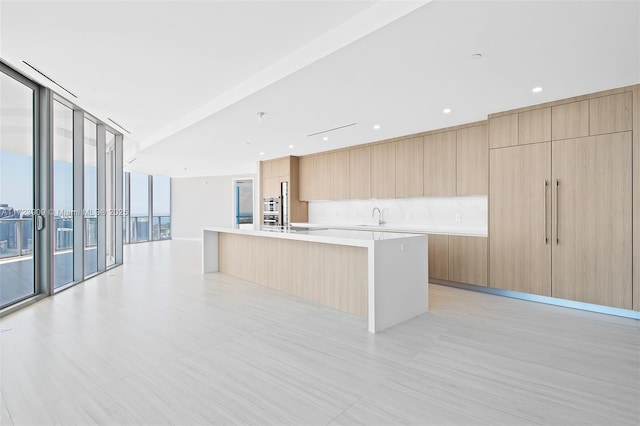 kitchen with sink, a wall of windows, an island with sink, and light brown cabinetry