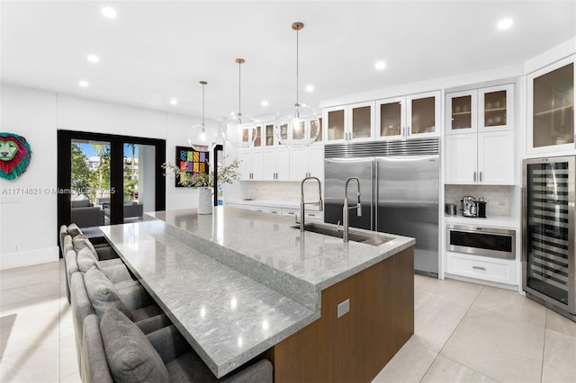 kitchen with light stone counters, a large island with sink, decorative light fixtures, white cabinets, and wine cooler