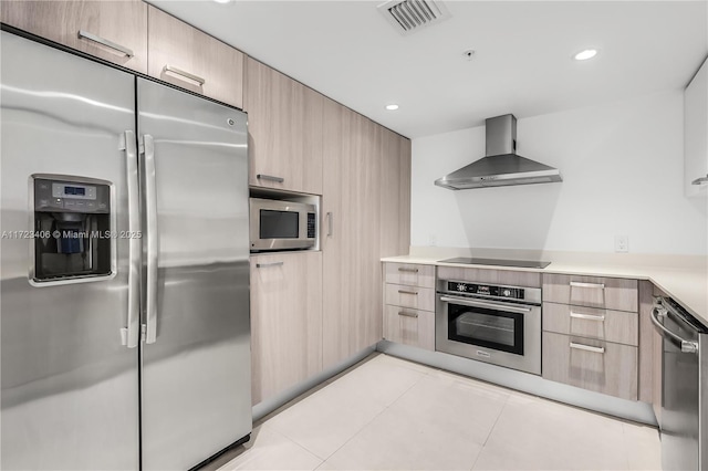 kitchen with light tile patterned flooring, appliances with stainless steel finishes, light brown cabinetry, and wall chimney range hood