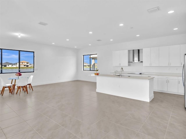 kitchen with wall chimney range hood, a center island with sink, light tile patterned floors, a healthy amount of sunlight, and white cabinets