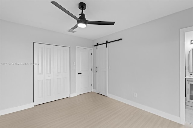 unfurnished bedroom with ensuite bath, ceiling fan, a barn door, light wood-type flooring, and a closet