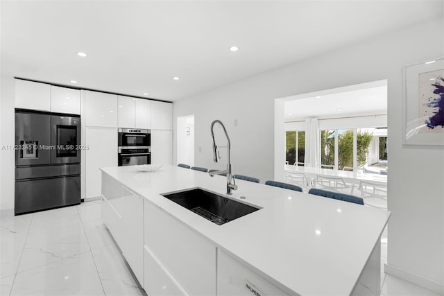 kitchen with stainless steel double oven, white cabinets, an island with sink, sink, and fridge with ice dispenser