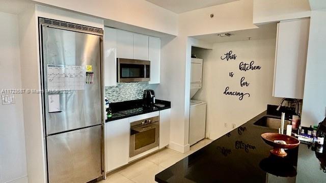 kitchen featuring stainless steel appliances, light tile patterned floors, stacked washer / dryer, decorative backsplash, and white cabinets