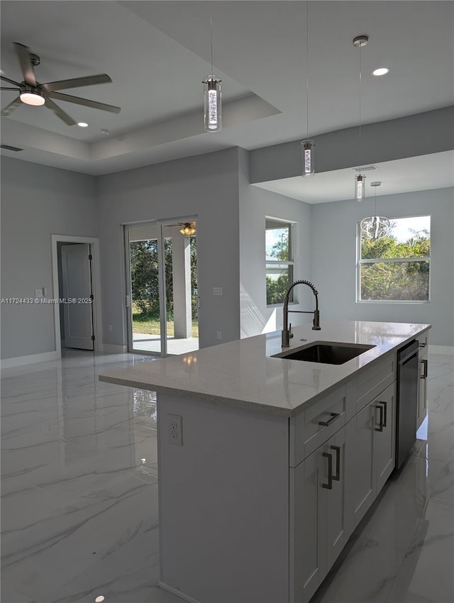 kitchen featuring light stone countertops, a tray ceiling, sink, decorative light fixtures, and a center island with sink