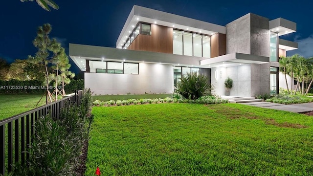 back of house at night with stucco siding, a yard, and fence