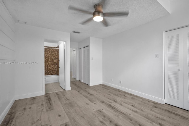 unfurnished bedroom featuring ensuite bathroom, ceiling fan, a textured ceiling, light hardwood / wood-style floors, and multiple closets