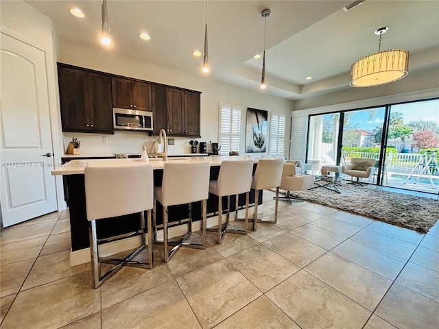 kitchen with a kitchen island with sink, a kitchen breakfast bar, a tray ceiling, decorative light fixtures, and dark brown cabinetry