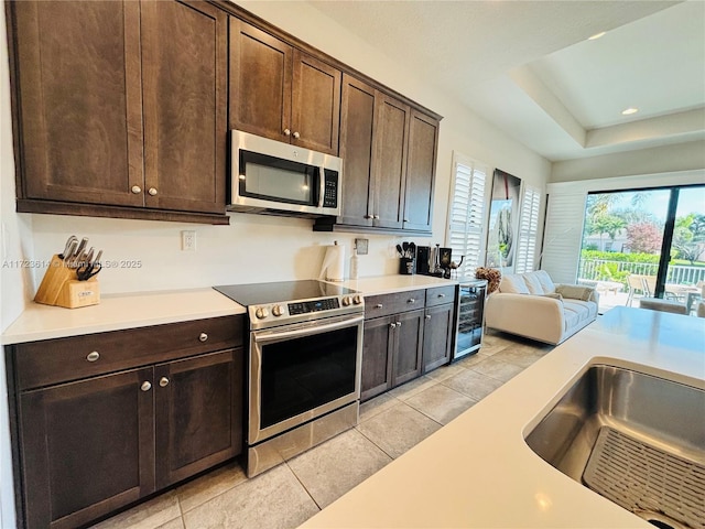 kitchen featuring beverage cooler, a raised ceiling, dark brown cabinets, light tile patterned floors, and appliances with stainless steel finishes