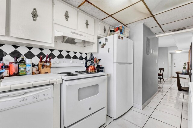 kitchen with white appliances, white cabinetry, a drop ceiling, light tile patterned floors, and under cabinet range hood
