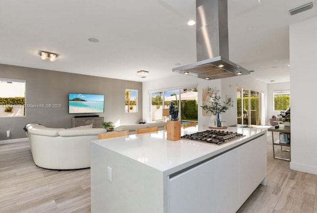 kitchen with island exhaust hood, light wood-type flooring, stainless steel gas stovetop, a kitchen island, and white cabinetry