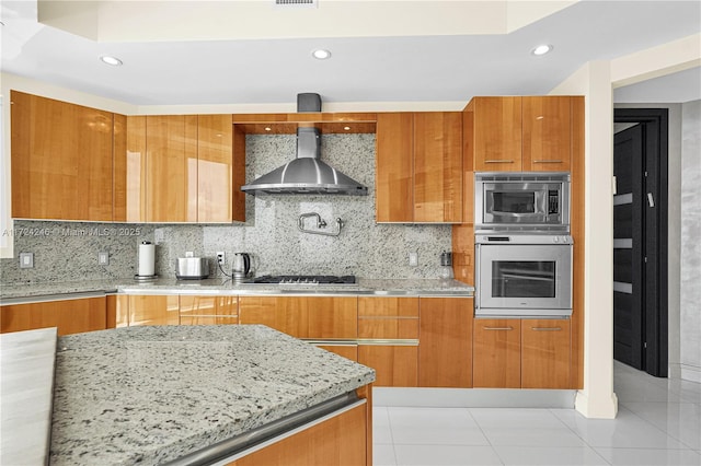 kitchen featuring light stone countertops, appliances with stainless steel finishes, wall chimney range hood, tasteful backsplash, and light tile patterned floors