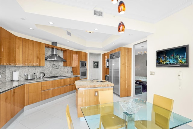 kitchen with backsplash, a kitchen island, a raised ceiling, built in appliances, and wall chimney exhaust hood