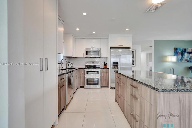 kitchen featuring stainless steel appliances, light tile patterned floors, tasteful backsplash, dark stone countertops, and white cabinets