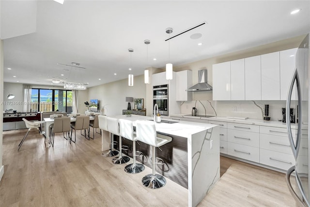 kitchen with appliances with stainless steel finishes, a kitchen island with sink, wall chimney range hood, white cabinets, and hanging light fixtures