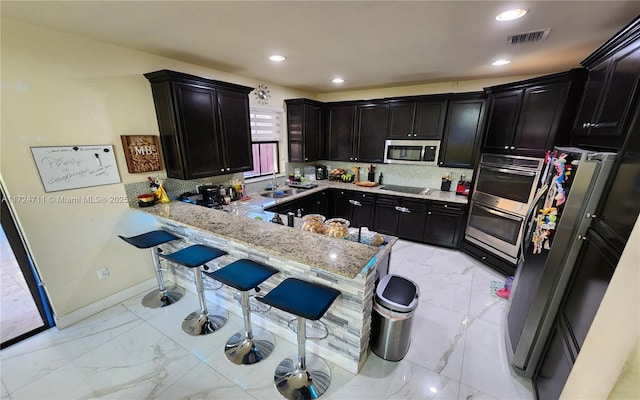 kitchen with light stone counters, backsplash, kitchen peninsula, a breakfast bar, and appliances with stainless steel finishes