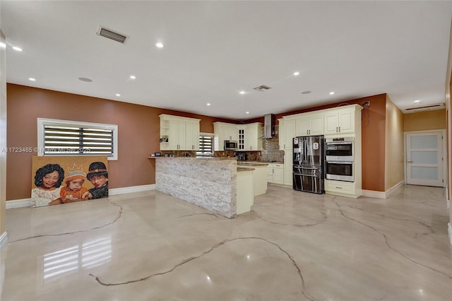 kitchen with wall chimney range hood, stone counters, appliances with stainless steel finishes, decorative backsplash, and kitchen peninsula