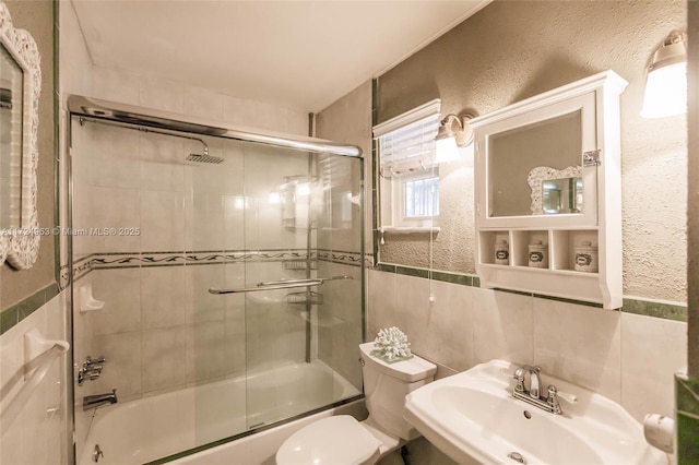 full bathroom with toilet, a sink, combined bath / shower with glass door, tile walls, and a textured wall