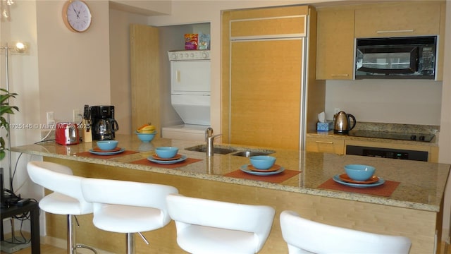 kitchen with stacked washing maching and dryer, black appliances, a breakfast bar, light stone counters, and sink