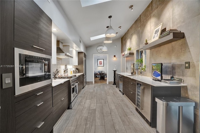 kitchen with decorative light fixtures, lofted ceiling with skylight, wall chimney range hood, sink, and appliances with stainless steel finishes