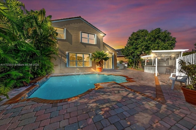 pool at dusk featuring a bar, a patio, pool water feature, and an outbuilding