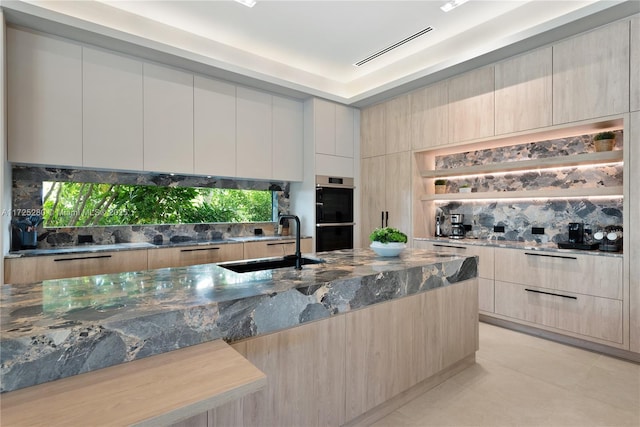 kitchen featuring sink, tasteful backsplash, light brown cabinetry, dark stone counters, and double oven