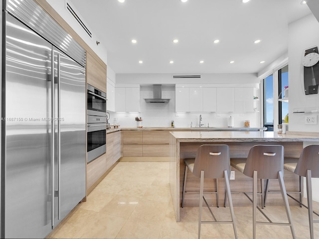 kitchen with appliances with stainless steel finishes, white cabinetry, a center island, wall chimney range hood, and a breakfast bar area