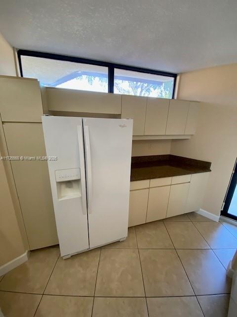 kitchen with a textured ceiling, white fridge with ice dispenser, light tile patterned flooring, and white cabinetry