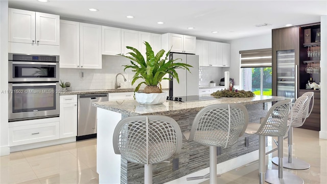 kitchen with stainless steel appliances, an island with sink, light stone countertops, light tile patterned floors, and white cabinetry