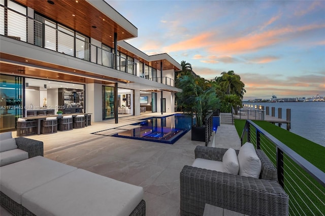 pool at dusk featuring a water view, outdoor lounge area, a lawn, and a patio area