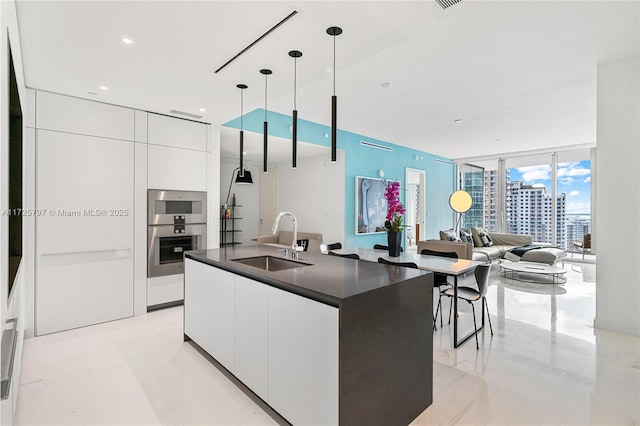 kitchen featuring decorative light fixtures, an island with sink, a wall of windows, white cabinetry, and sink