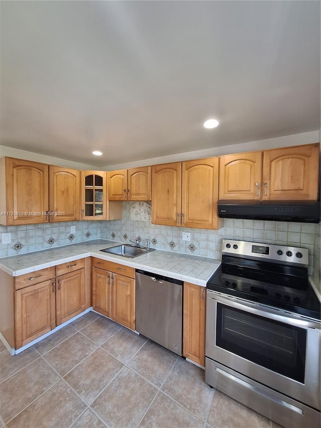 kitchen featuring tile counters, decorative backsplash, stainless steel appliances, under cabinet range hood, and a sink