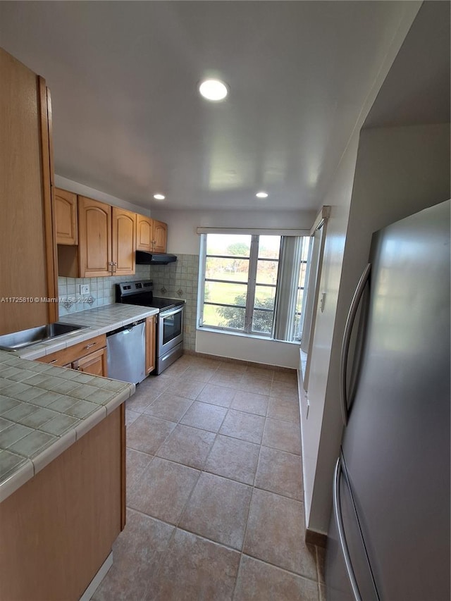 kitchen with light tile patterned flooring, under cabinet range hood, stainless steel appliances, tile counters, and decorative backsplash