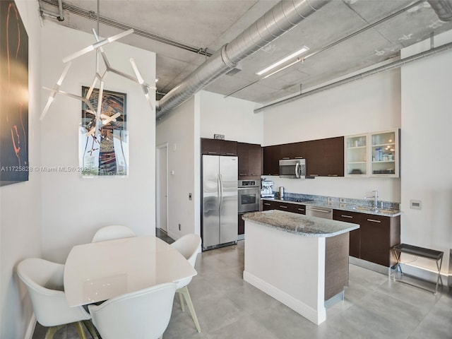 kitchen featuring light stone counters, a center island, a high ceiling, appliances with stainless steel finishes, and dark brown cabinets