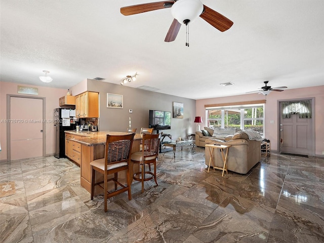 kitchen with light brown cabinetry, black fridge, kitchen peninsula, ceiling fan, and a breakfast bar