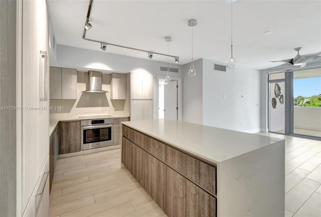 kitchen featuring stainless steel oven, wall chimney exhaust hood, ceiling fan, a spacious island, and rail lighting