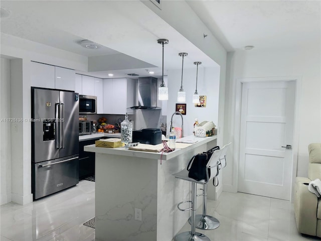 kitchen featuring stainless steel appliances, white cabinets, a kitchen bar, hanging light fixtures, and wall chimney range hood