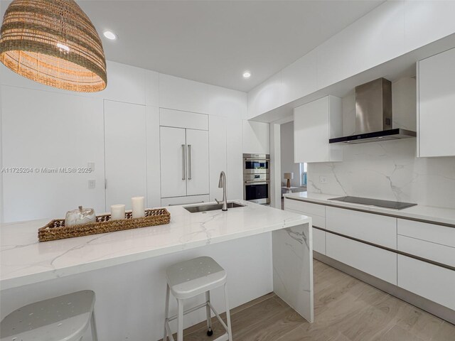 kitchen with pendant lighting, wall chimney range hood, black electric stovetop, white cabinets, and stainless steel double oven