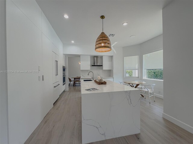 kitchen featuring white cabinetry, wall chimney range hood, decorative light fixtures, and a large island with sink