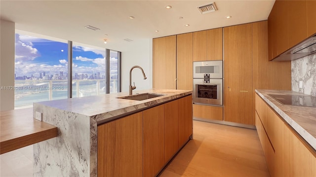 kitchen featuring a kitchen island with sink, black electric cooktop, stainless steel double oven, sink, and light hardwood / wood-style flooring