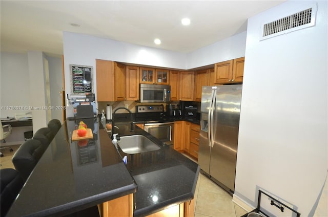 kitchen featuring stainless steel appliances, a peninsula, a sink, brown cabinetry, and dark countertops