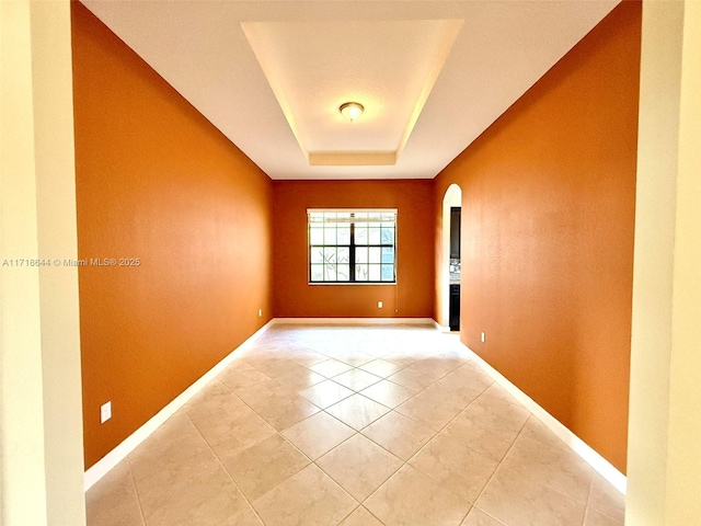 empty room featuring a tray ceiling, light tile patterned flooring, baseboards, and arched walkways