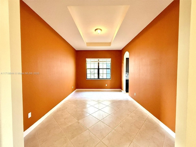 empty room with arched walkways, a raised ceiling, baseboards, and light tile patterned flooring