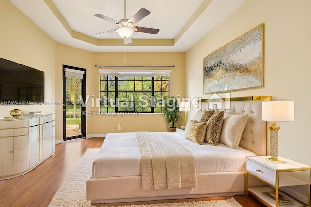 bedroom with a ceiling fan, wood finished floors, a raised ceiling, baseboards, and access to exterior