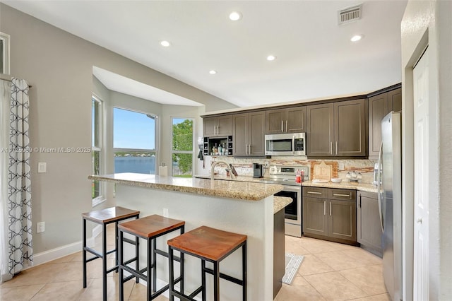 kitchen featuring stainless steel appliances, a water view, decorative backsplash, light stone countertops, and a center island with sink