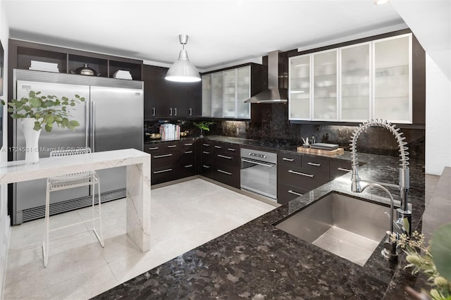 kitchen featuring hanging light fixtures, appliances with stainless steel finishes, wall chimney exhaust hood, sink, and dark brown cabinetry